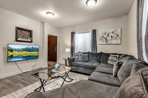 Una sala de estar con un televisor de pantalla plana grande, un sofá gris, una mesa de centro de vidrio y un elemento decorativo en la pared.