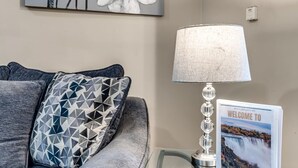 Ein Wohnzimmer mit grauem Sofa, einem geometrisch gemusterten Kissen, einer Wandlampe und einem gerahmten Willkommensschild.