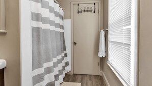 Ein Badezimmer mit Duschvorhang, Handtuchhalter, einer Tür und einem Fenster mit Jalousien.
