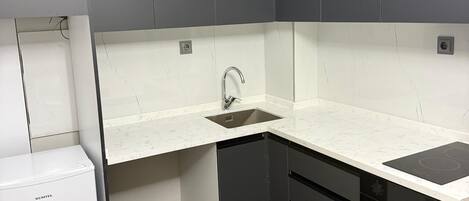 Cocina compacta con gabinetes grises, encimera blanca, lavavajillas integrado y refrigerador blanco.