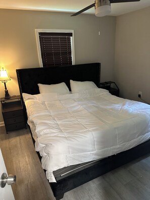 Un lit bien fait, avec une literie blanche, une tête de lit sombre, une table de chevet avec une lampe et un ventilateur de plafond.