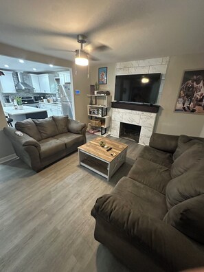 Ein Wohnzimmer mit Kamin, Fernseher und einer Küche im Hintergrund.