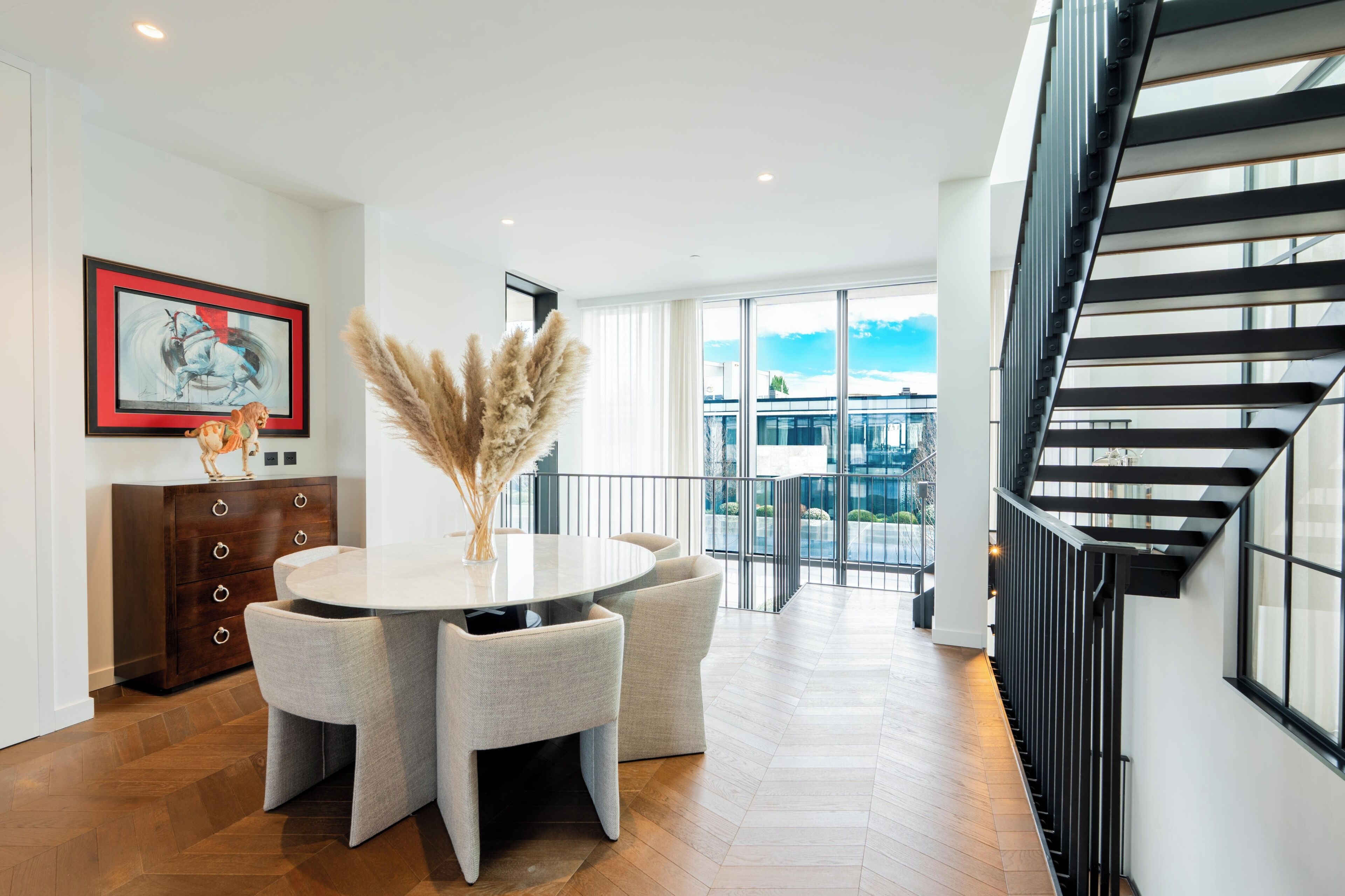 A modern interior with a round dining table, chairs, a wooden cabinet, a framed artwork, and a staircase.