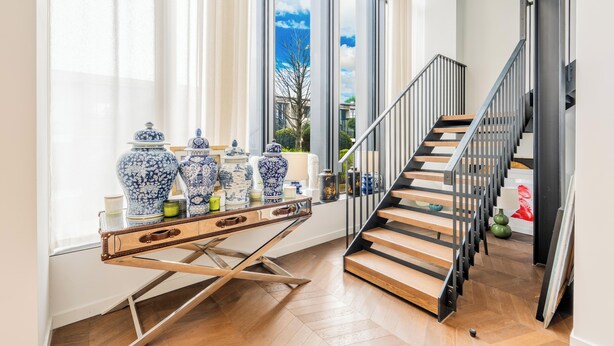A staircase with wooden steps and metal railings, a table with blue and white vases, and a view of the sky through large windows. - Luxury 4-Bed Duplex Penthouse (London)