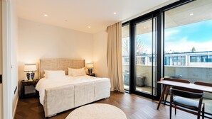 A bedroom with a large bed, bedside lamps, a round rug, a dining table with chairs, and a view through a sliding glass door. - Luxury 4-Bed Duplex Penthouse (London)