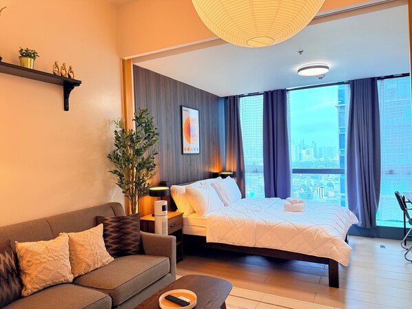 Una habitación de hotel moderna con cama, sofá, mesita de noche, lámpara, planta en maceta y vistas a la ciudad.