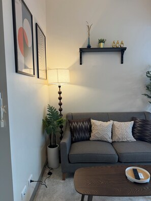 Una sala de estar con un sofá gris, almohadones decorativos, una lámpara de pie, una mesita con un control remoto y un estante en la pared con una planta y figuritas.