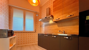 A modern kitchen with a microwave, stove, and sink, wooden cabinets, and a window with blinds. - Poetto Beach | 5 Min Walk [WiFi & AC] (Cagliari)
