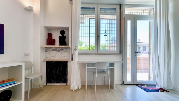 A room with a white desk, a chair, a fireplace, and a view through a window with metal bars. - Poetto Beach | 5 Min Walk [WiFi & AC] (Cagliari)