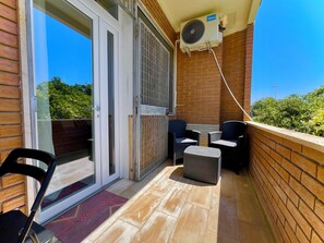 Ein Balkon mit Schiebeglastür, zwei Korbstühle, einem kleinen Tisch, einem Fußhocker aus Korbgeflecht und einem Deckenventilator.