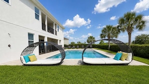 Un espace aménagé au bord de la piscine, avec deux fauteuils en osier, une piscine et un bâtiment blanc doté de balcons.