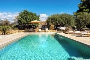 Ein rechteckiger Pool mit klarem, blauem Wasser, umgeben von Liegestühlen und Sonnenschirmen, vor einer Kulisse aus Bäumen und einer Steinmauer.