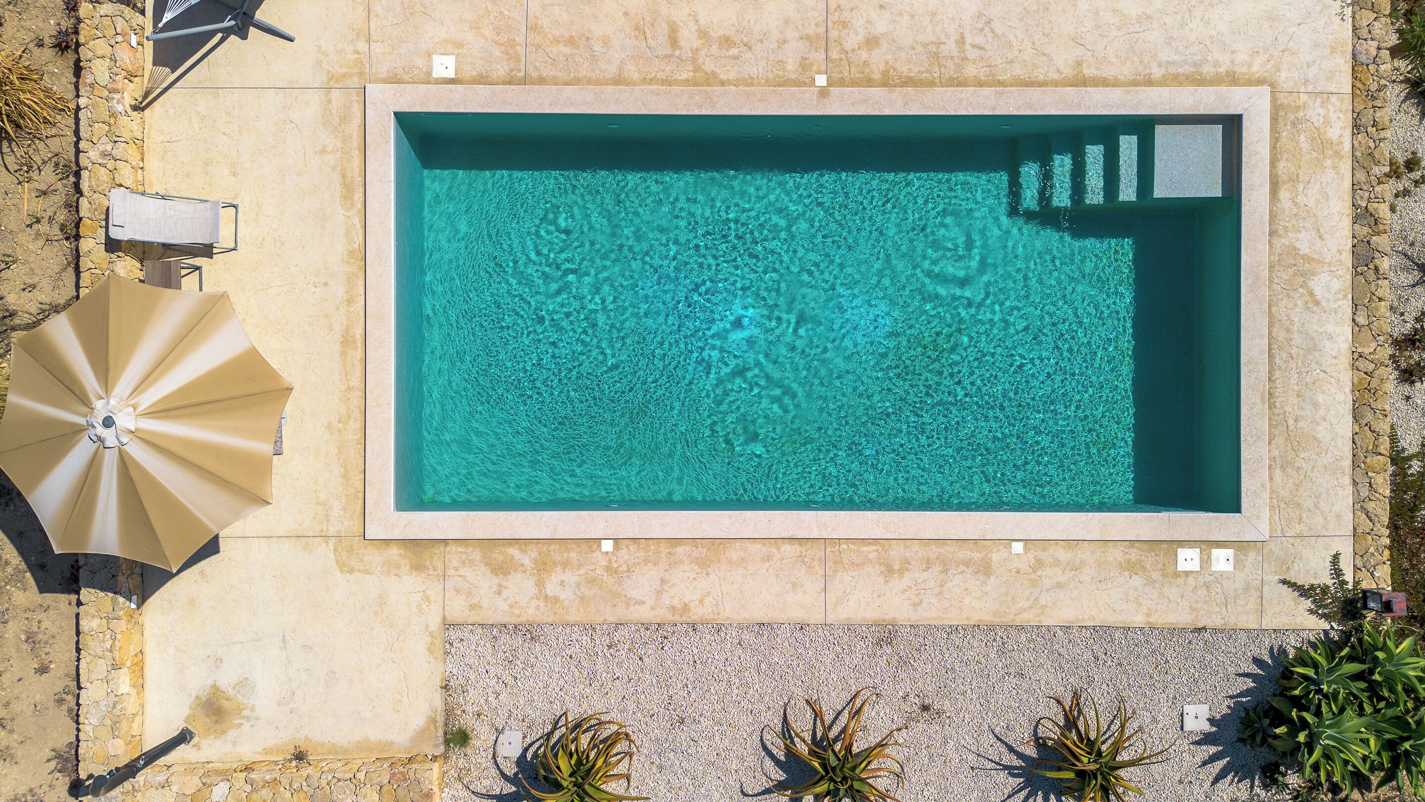 Una piscina rectangular de aguas cristalinas, rodeada por un solárium de piedra y algunas sombrillas.