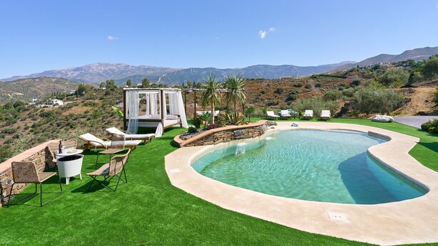 Una piscina de borde curvo, rodeada de césped artificial, muebles de mimbre y un muro de piedra.