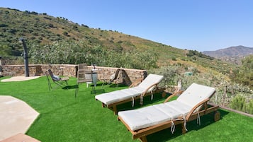 Een terras met twee ligbedden, een tafel en stoelen, omgeven door een stenen muur en uitzicht op een glooiend landschap.