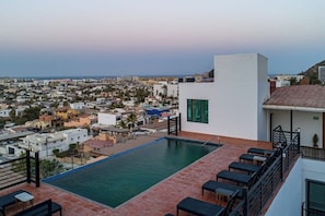 Une piscine sur le toit offrant une vue sur la ville, un immeuble blanc doté d’un balcon et une terrasse avec des sièges.