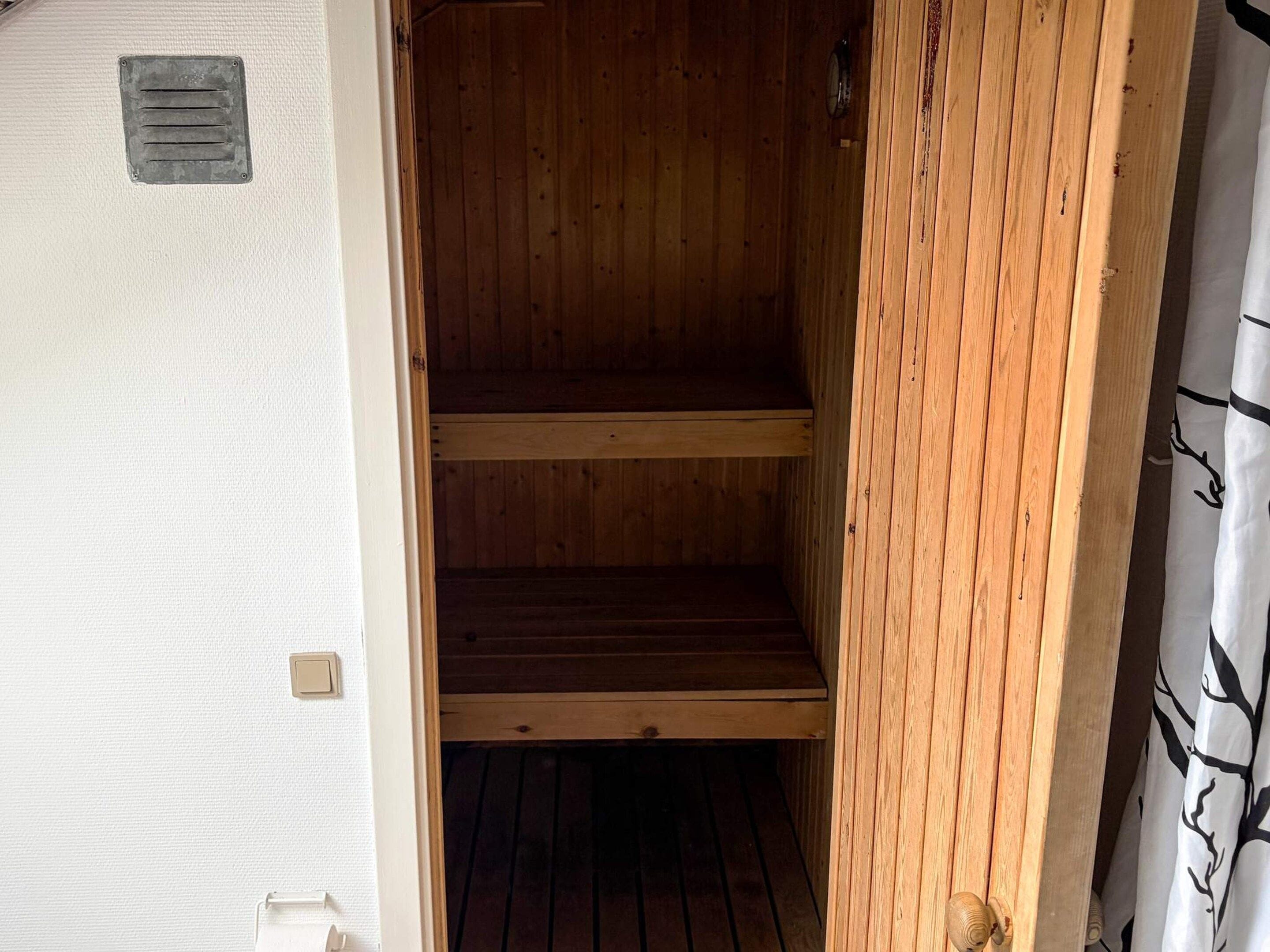 Un intérieur en bois, avec une grille de ventilation en métal et une porte donnant sur une pièce dont la fenêtre est ornée d’un rideau à motifs.