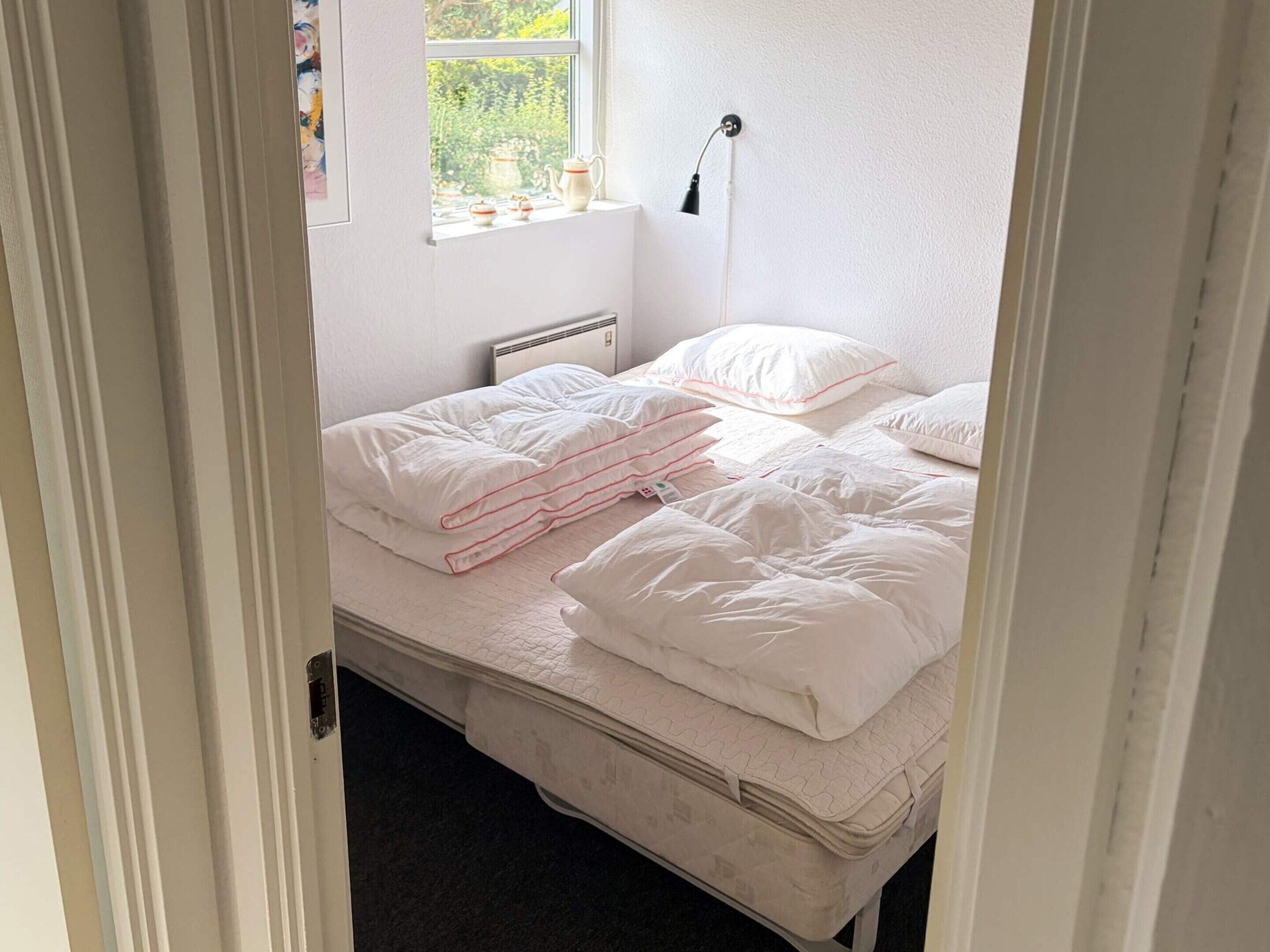 Quarto com cama, almofadas, abajur, janela com vista para área verde e radiador.