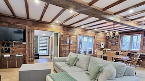 A spacious living room with a large sofa, a dining area with wooden furniture, and a kitchen visible through an open doorway. - Le Nordique | Private Sauna & Spa | Lake access (Val-des-Lacs)