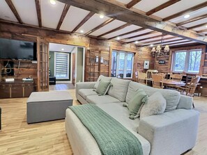 A spacious living room with a large sofa, a dining area with wooden furniture, and a kitchen visible through an open doorway. - Le Nordique | Private Sauna & Spa | Lake access (Val-des-Lacs)
