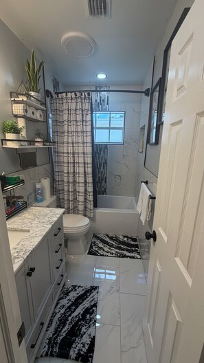 A bathroom with a toilet, bathtub, and shower curtain. There are shelves with toiletries and a potted plant. - COZY - CLEAN QUITE ROOM NEAR HARD ROCK - The Radiance Room🏡 (Miramar)