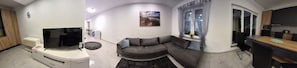 Een woonkamer met een flatscreen televisie, een grijze bank, een eethoek met een houten tafel en een keuken met moderne apparatuur.