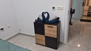 A room with a bed, a small wooden and black cabinet, a wall socket, and a door leading to another room.