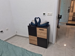 Ein Zimmer mit einem Bett, einem kleinen Holzschrank in Schwarz, einer Steckdose und einer Tür zu einem anderen Raum.