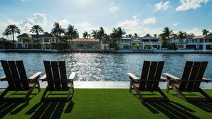 Uitzicht op het water met drie houten stoelen, een gazon en een rij huizen langs de waterlijn.