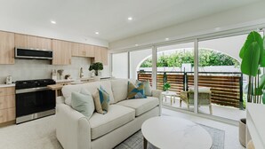 Een moderne keuken met een ingebouwde oven en magnetron, een beige bank met decoratieve kussens, een ronde eettafel en een grote schuifdeur naar een buitenruimte.