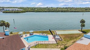 Ein Schwimmbecken mit Liegestühlen, ein Tennisplatz und ein Blick auf ein Gewässer mit einer entfernten Küstenlinie.