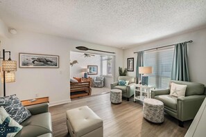 Ein großzügiges Wohnzimmer mit einer Couch, Hocker und Sesseln, einem Deckenventilator und Blick auf das angrenzende Schlafzimmer.
