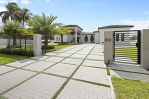 Una entrada con portón a una zona residencial, con camino de entrada pavimentado, palmeras y una casa con el número 80.