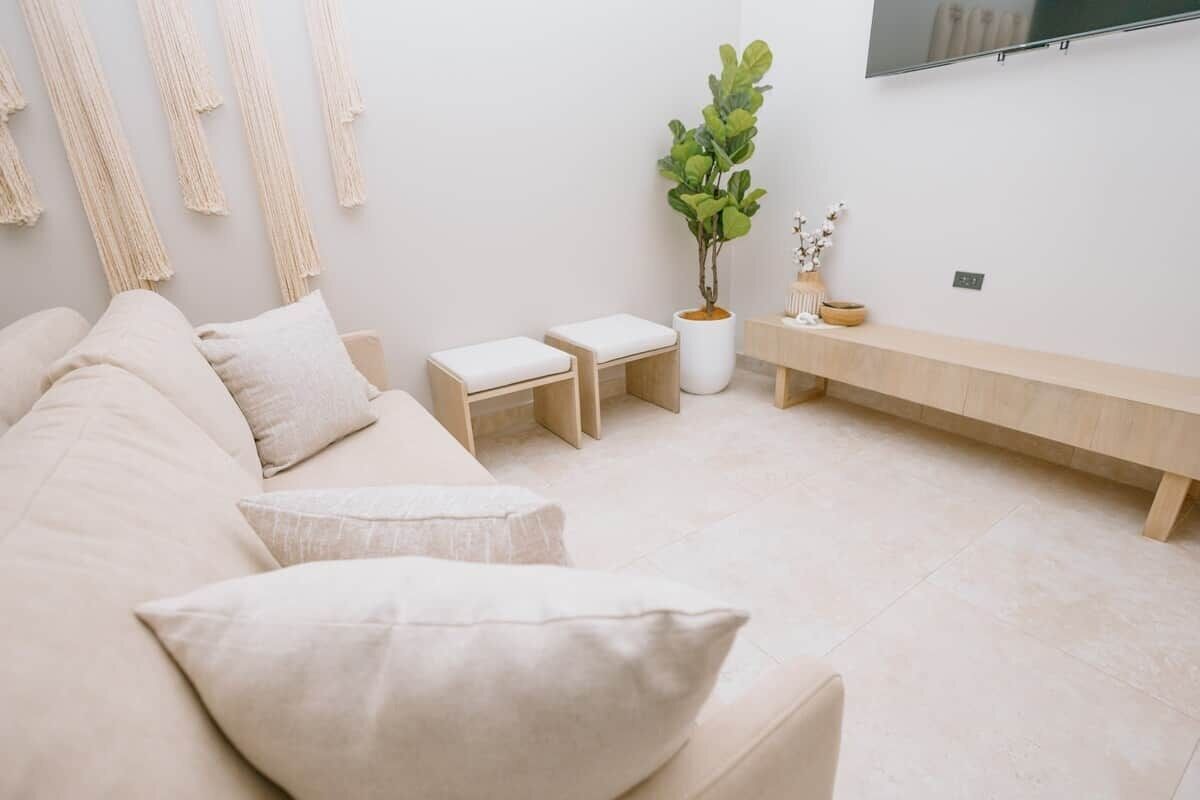 Un salon avec un canapé beige, deux poufs blancs, un banc en bois, une plante en pot et un téléviseur fixé au mur.