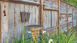 Una estructura rústica de madera con un letrero de metal con la letra 'W', un bebedero para pájaros y follaje circundante.