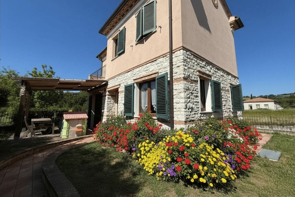 A two-story stone house with a covered patio, a flower garden, and a clear blue sky.