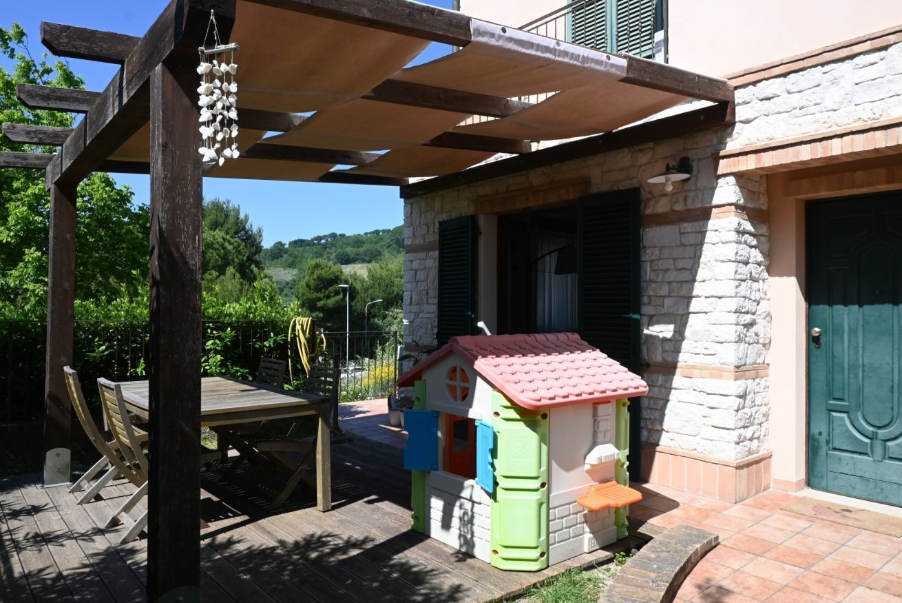 A wooden pergola, a children's playhouse, a stone building with a green door, and a patio table with chairs.