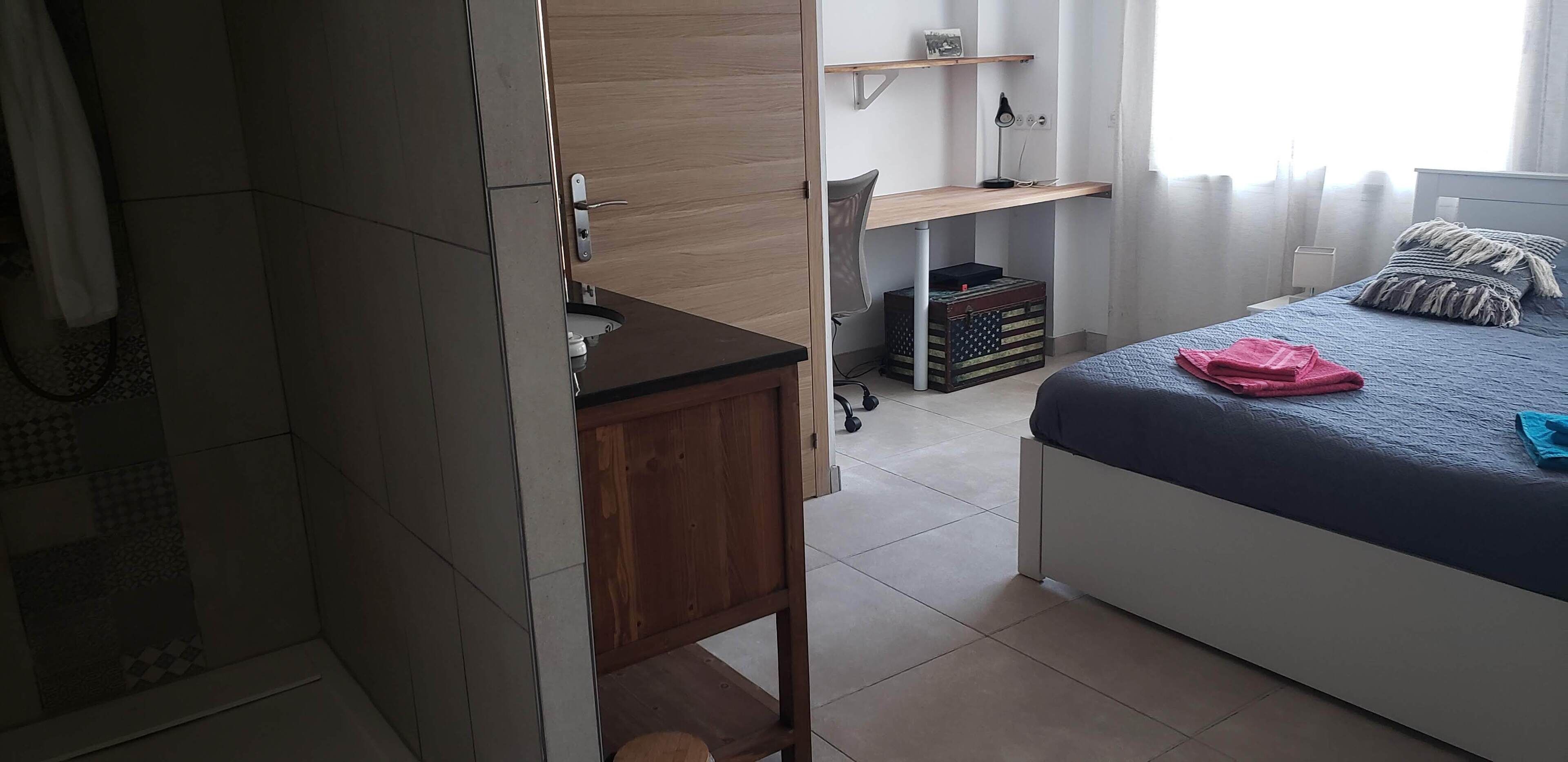 A bedroom with a bed, a desk, a chair, a wardrobe, and a small storage unit.