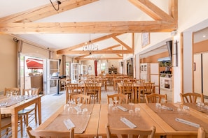 A spacious dining area with wooden tables and chairs, a chandelier, and a view of the kitchen area. - Ardeche House With Spacious Terrace for 8 (Grospierres)
