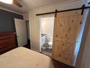 Quarto com cama, cabeceira de madeira, porta de estilo rústico deslizante e vista para um banheiro com vaso sanitário e pia.