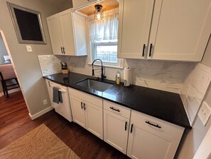 Cozinha com armários brancos, bancada preta, janela com cortinas e piso de madeira.