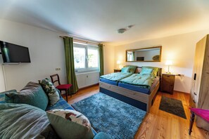 Ein Schlafzimmer mit einem Bett, einem Fernseher, einem Fenster mit Vorhängen, einem Nachttisch und einem Sessel.