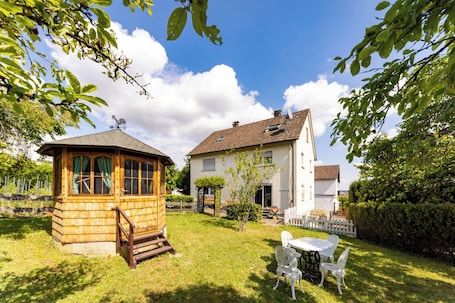 A wooden gazebo, a white house with a tiled roof, and a white table with chairs in a grassy yard.. Apartment Marie