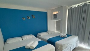 A hotel room with two beds, a blue accent wall, a window with curtains, and an air conditioning unit. - Anchor Flats Condominium by the Sea (Ipojuca)