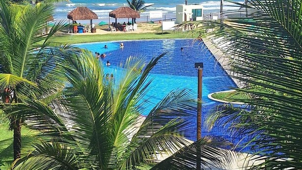 A pool area with a view of the ocean, palm trees, and a thatched-roof structure. - Anchor Flats Condominium by the Sea (Ipojuca)