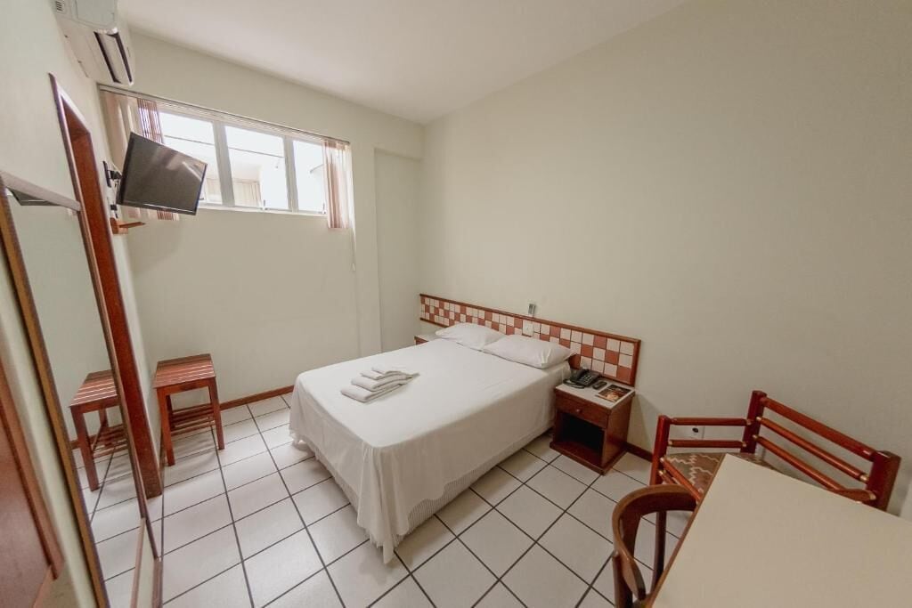 Una cama individual con cabecera de madera, una mesita de noche con lámpara, una silla y un televisor fijado a la pared.