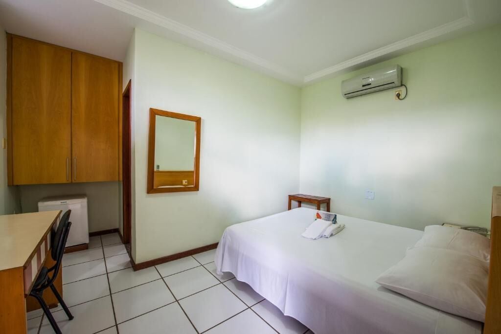 Una habitación de hotel con cama, escritorio, silla, mesita, espejo y aire acondicionado.