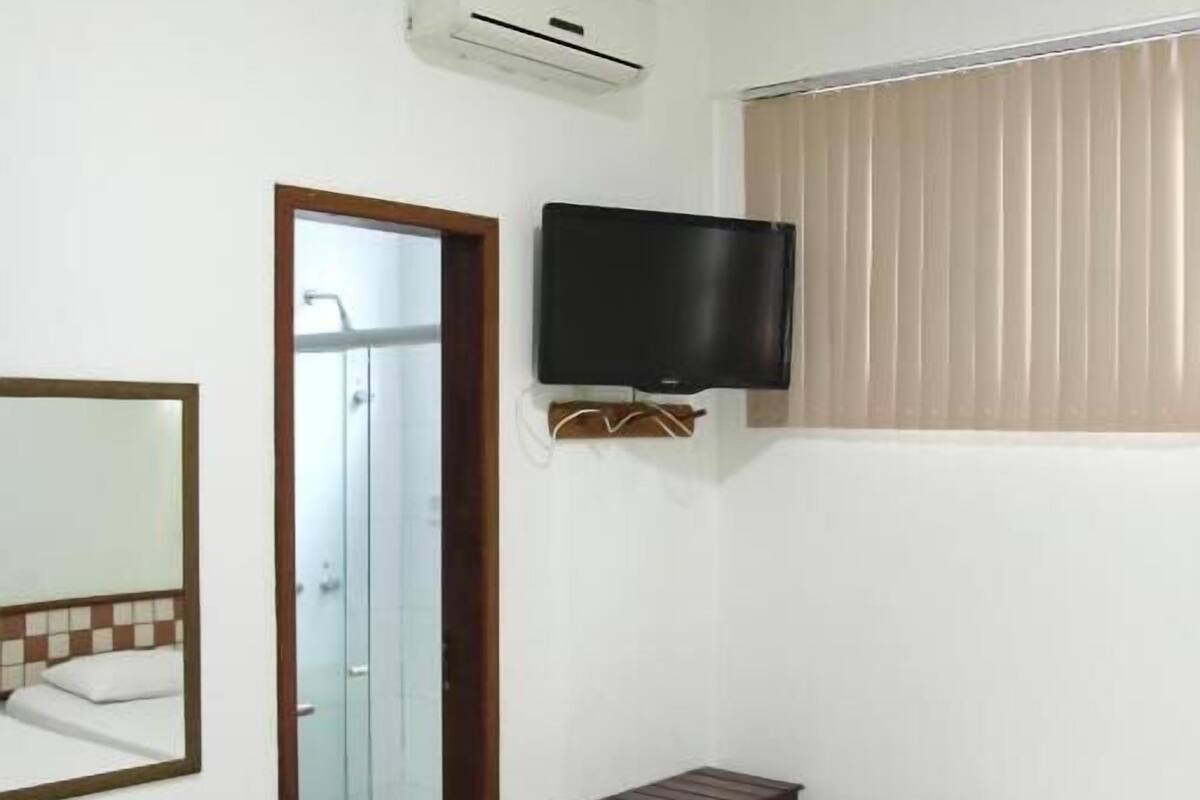 A hotel room with a bed, a television mounted on the wall, a small table, a mirror, and an air conditioning unit.
