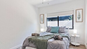 Une chambre à coucher avec un lit, une fenêtre avec des stores, une table de chevet et un tableau encadré au mur.