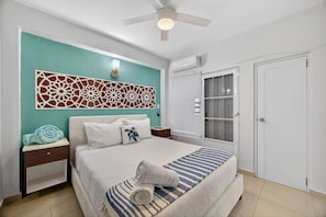 Ein Schlafzimmer mit Bett, Nachttischen, Deckenventilator, dekorativer Wandverkleidung, einem Fenster mit Jalousien und einer Tür.